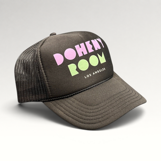 Raree Finds Doheny Room black mesh trucker hat with pastel logo on a structured foam front panel and curved brim.