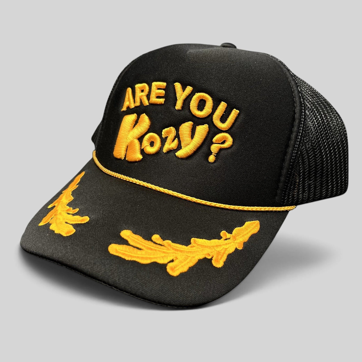 Raree Finds Are You Kozy? black trucker hat with gold raised embroidery, gold rope detail, and leaf bill accents.