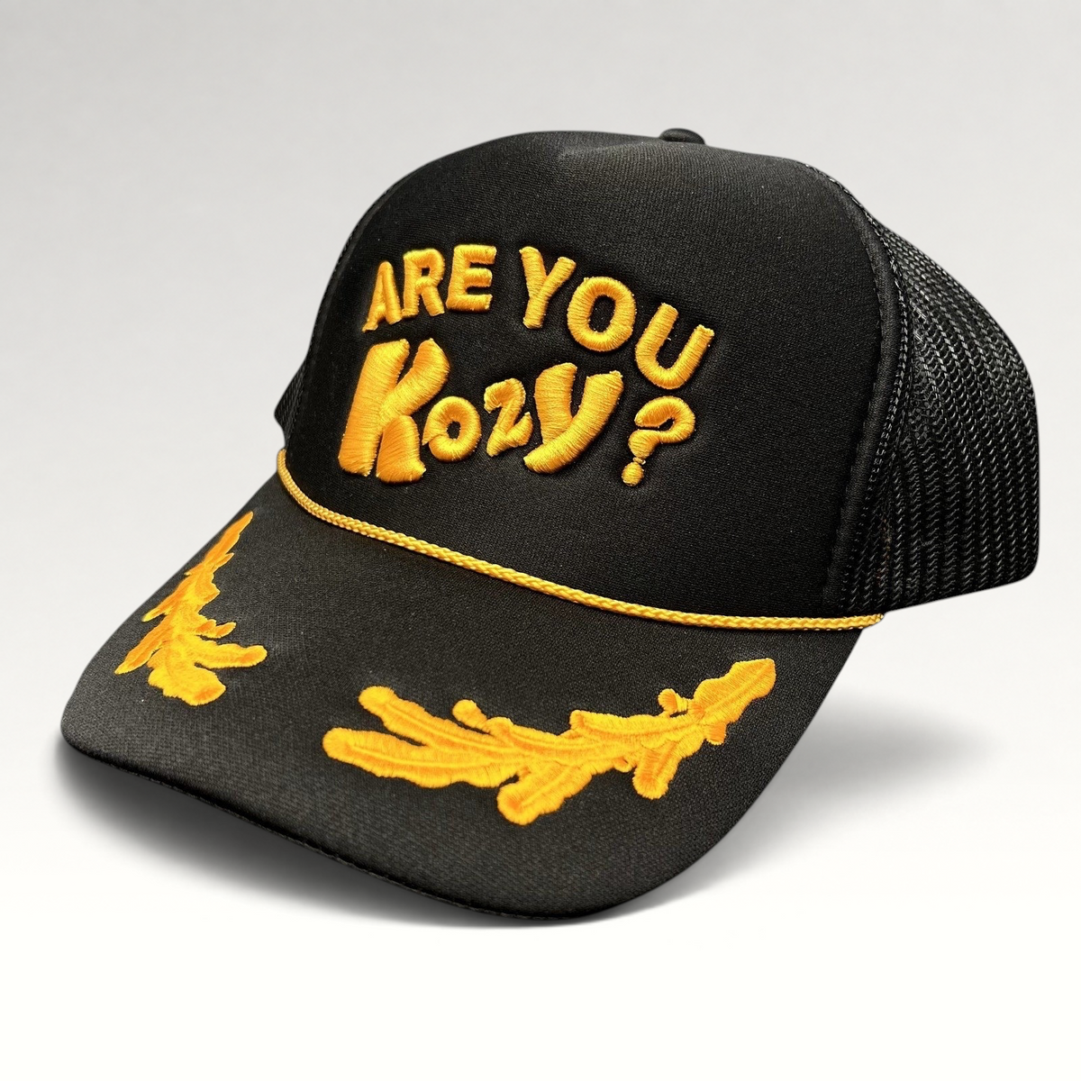 Raree Finds Are You Kozy? black trucker hat with gold raised embroidery, gold rope detail, and leaf bill accents.