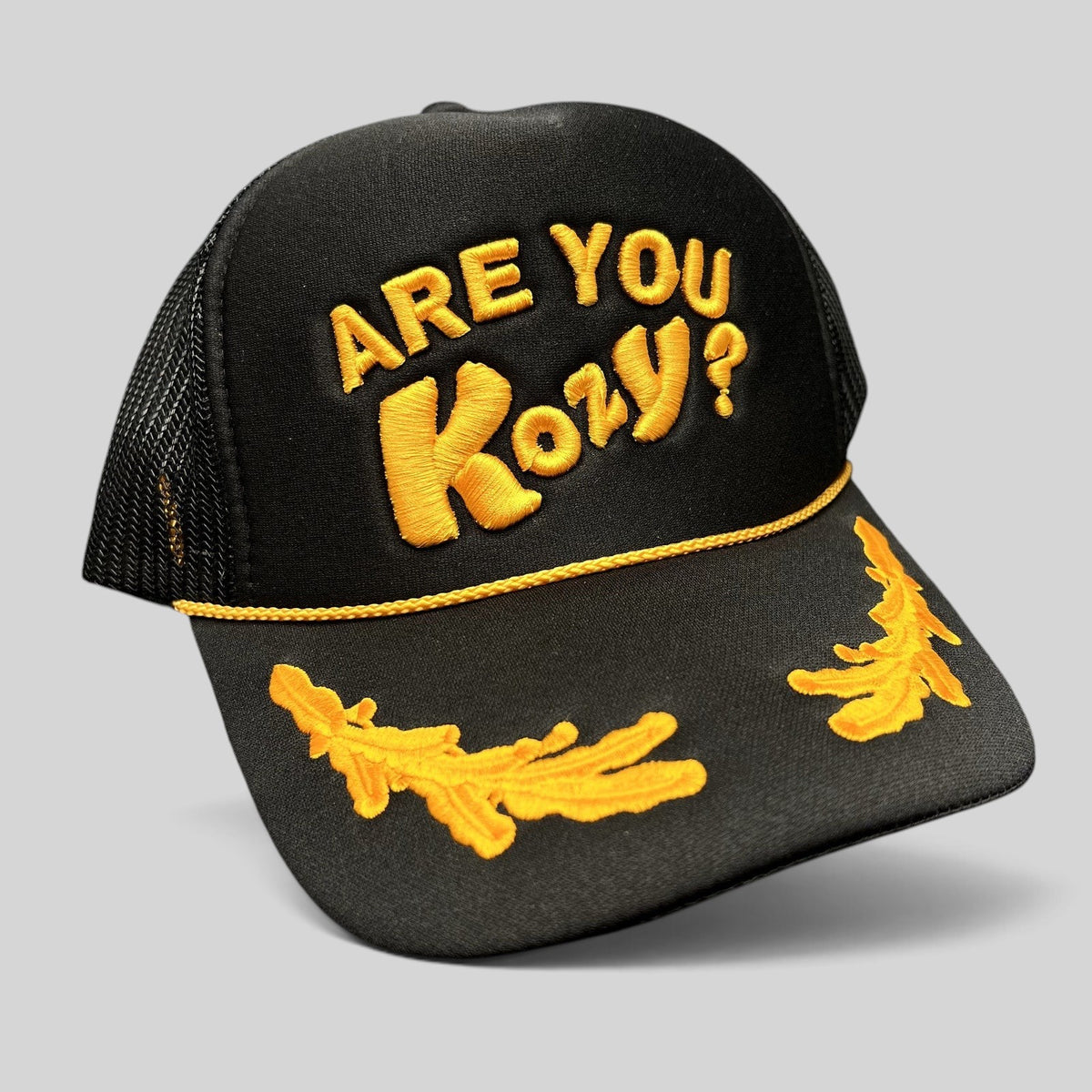 Raree Finds Are You Kozy? black trucker hat with gold raised embroidery, gold rope detail, and leaf bill accents.