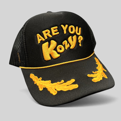 Raree Finds Are You Kozy? black trucker hat with gold raised embroidery, gold rope detail, and leaf bill accents.