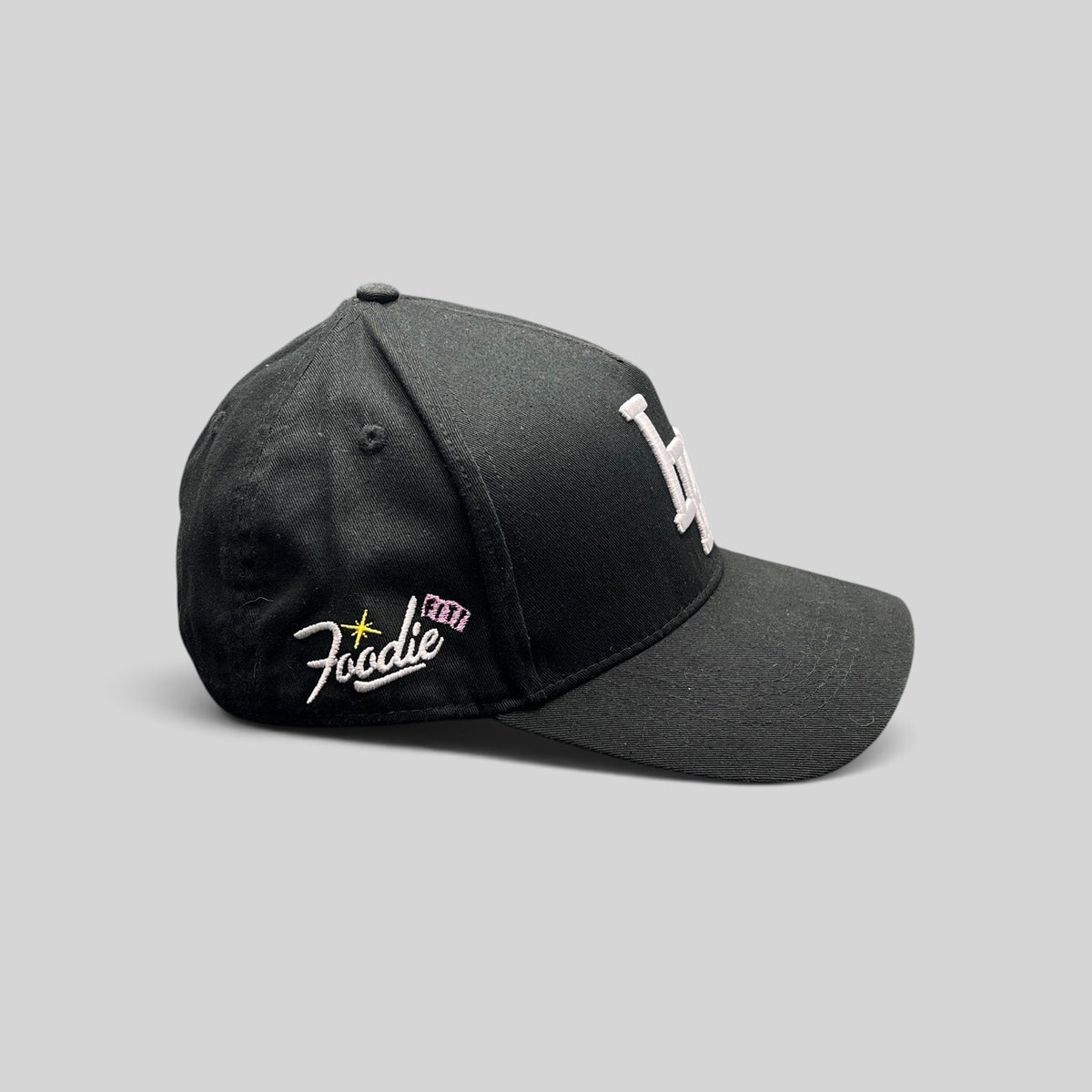 Raree Finds black Okami Brand strapback hat with white embroidered LV logo and side Foodie script detail