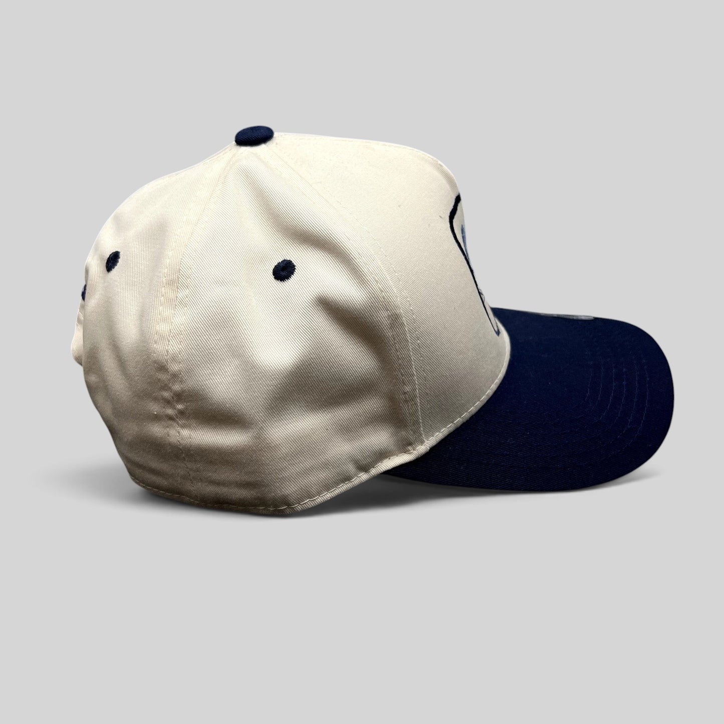 Raree Finds The Flying Cock NYC snapback hat with a cream crown and navy blue brim, side profile view.