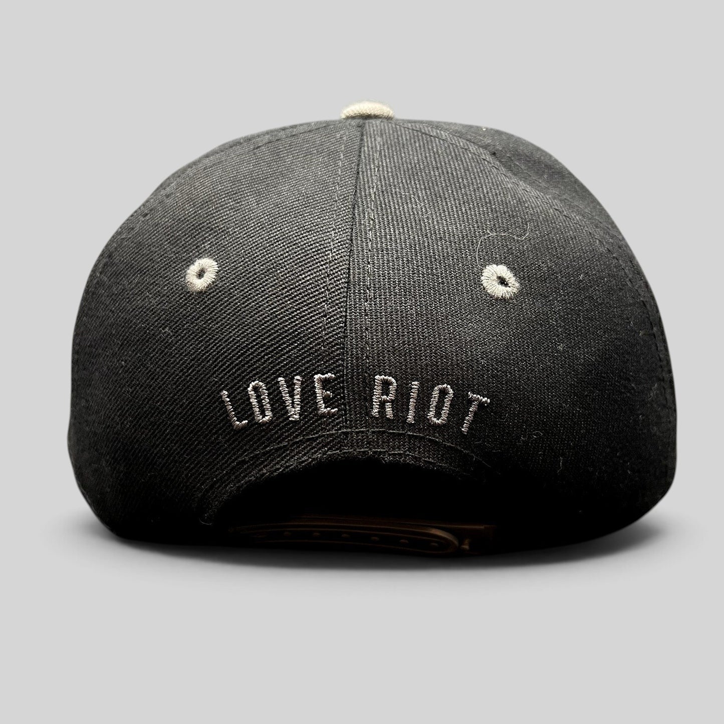 Raree Finds black snapback hat featuring embroidered Love Riot script on the back and gray eyelet details.