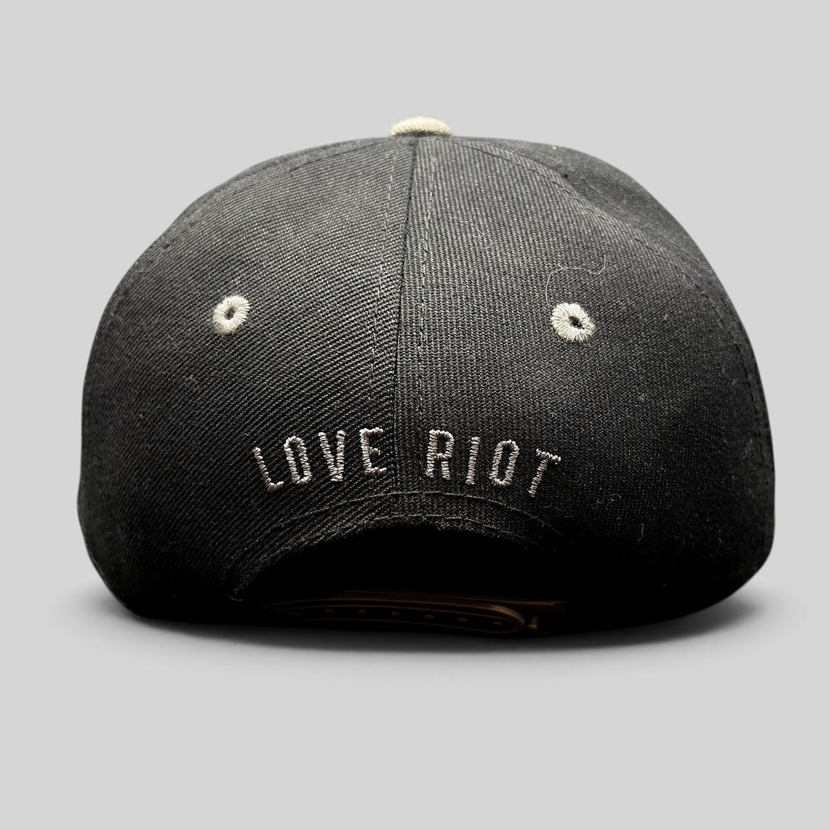 Raree Finds black snapback hat featuring embroidered Love Riot script on the back and gray eyelet details.
