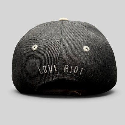Raree Finds black snapback hat featuring embroidered Love Riot script on the back and gray eyelet details.