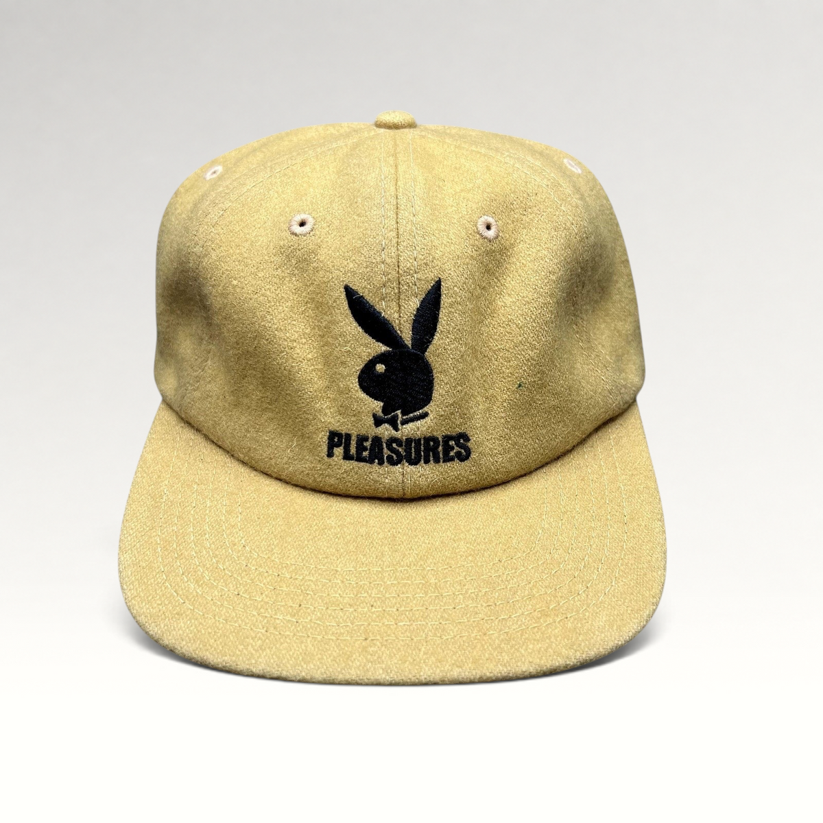 Raree Finds mustard wool strapback hat featuring an embroidered black Playboy Bunny and PLEASURES logo on the front.