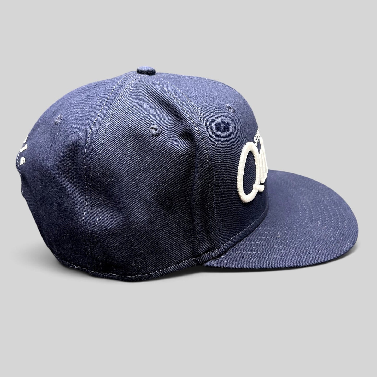 Raree Finds navy blue Quixote Studios snapback hat with raised white embroidery and flat brim on a dark background.