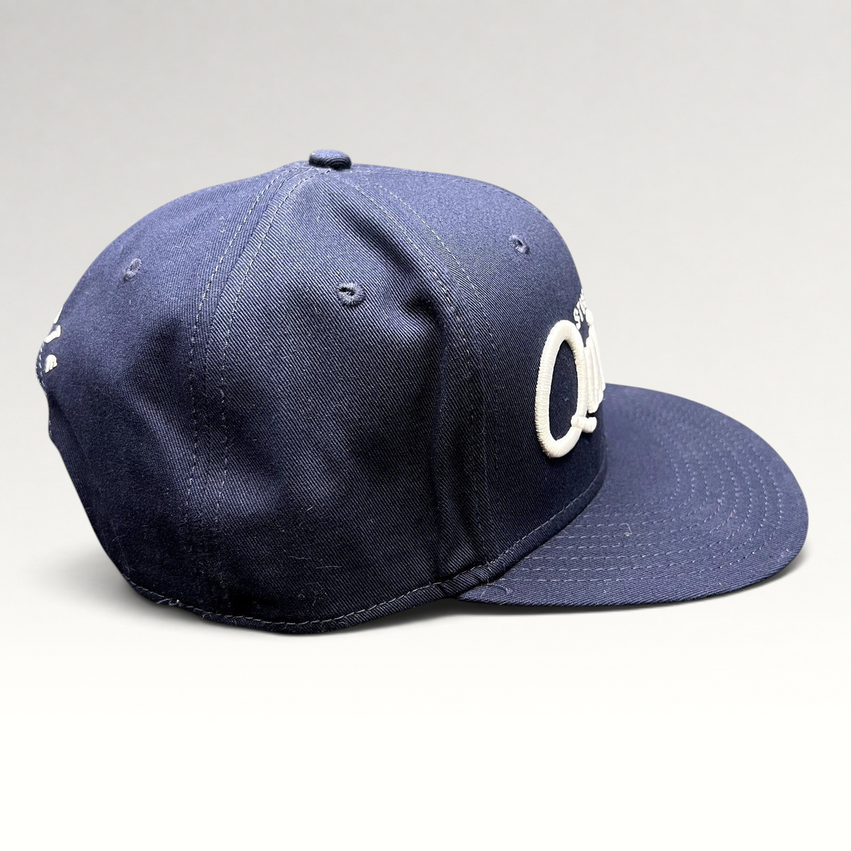 Raree Finds navy blue Quixote Studios snapback hat with raised white embroidery and flat brim on a dark background.