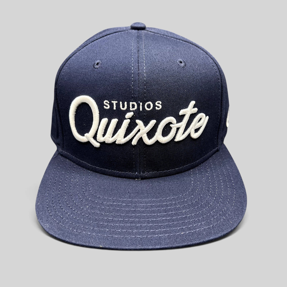 Raree Finds navy blue snapback hat with raised white Quixote Studios embroidery on the structured crown and flat brim.