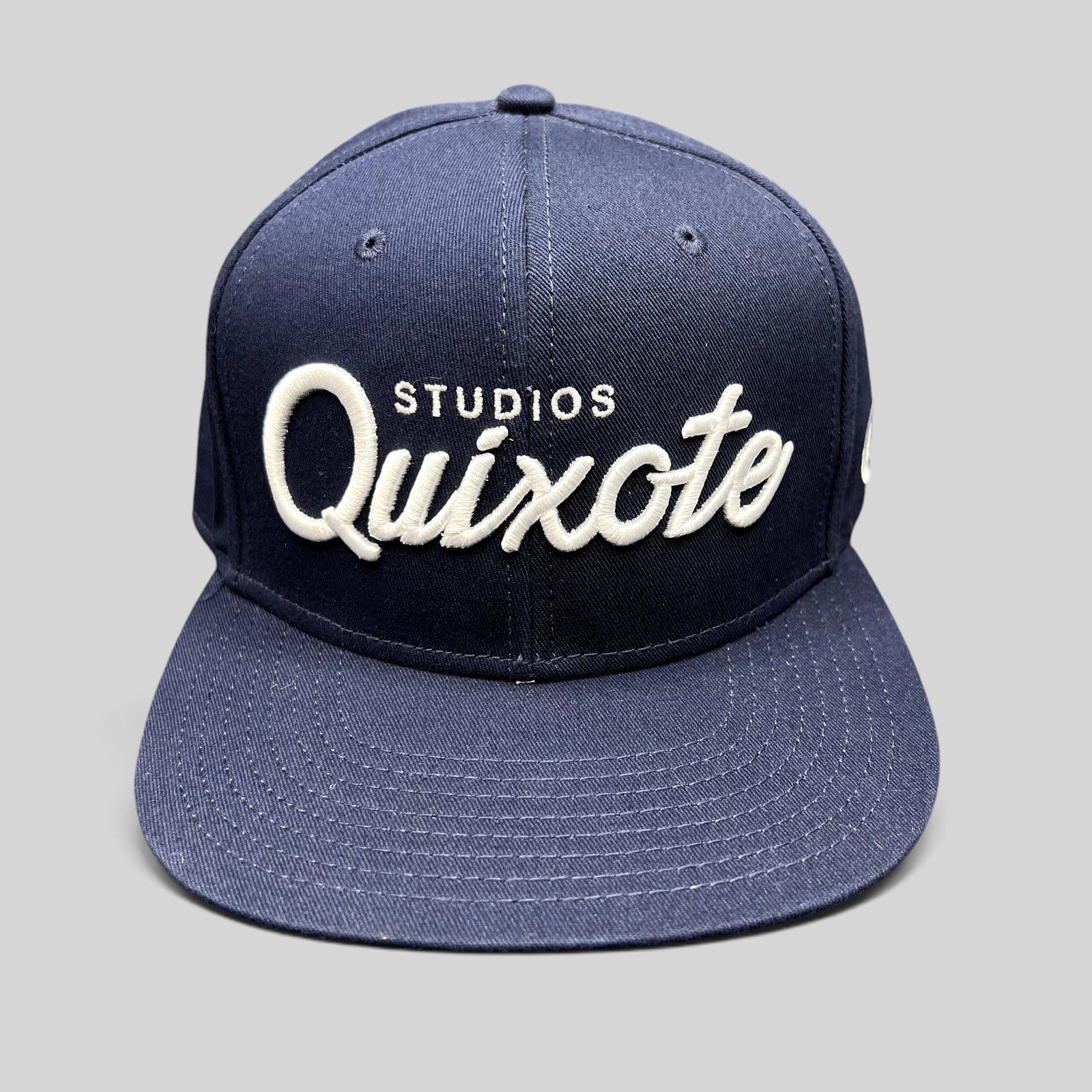 Raree Finds navy blue snapback hat with raised white Quixote Studios embroidery on the structured crown and flat brim.