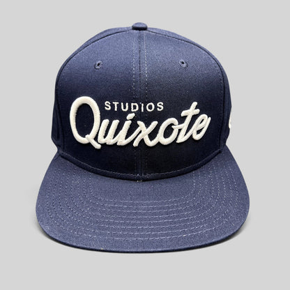 Raree Finds navy blue snapback hat with raised white Quixote Studios embroidery on the structured crown and flat brim.