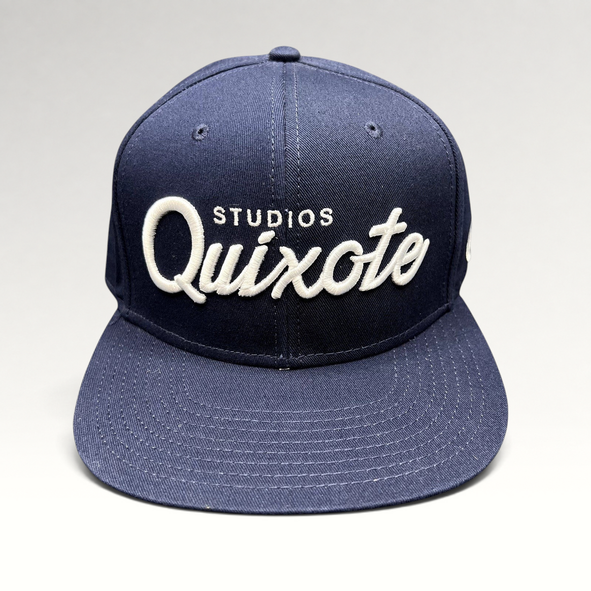 Raree Finds navy blue snapback hat with raised white Quixote Studios embroidery on the structured crown and flat brim.