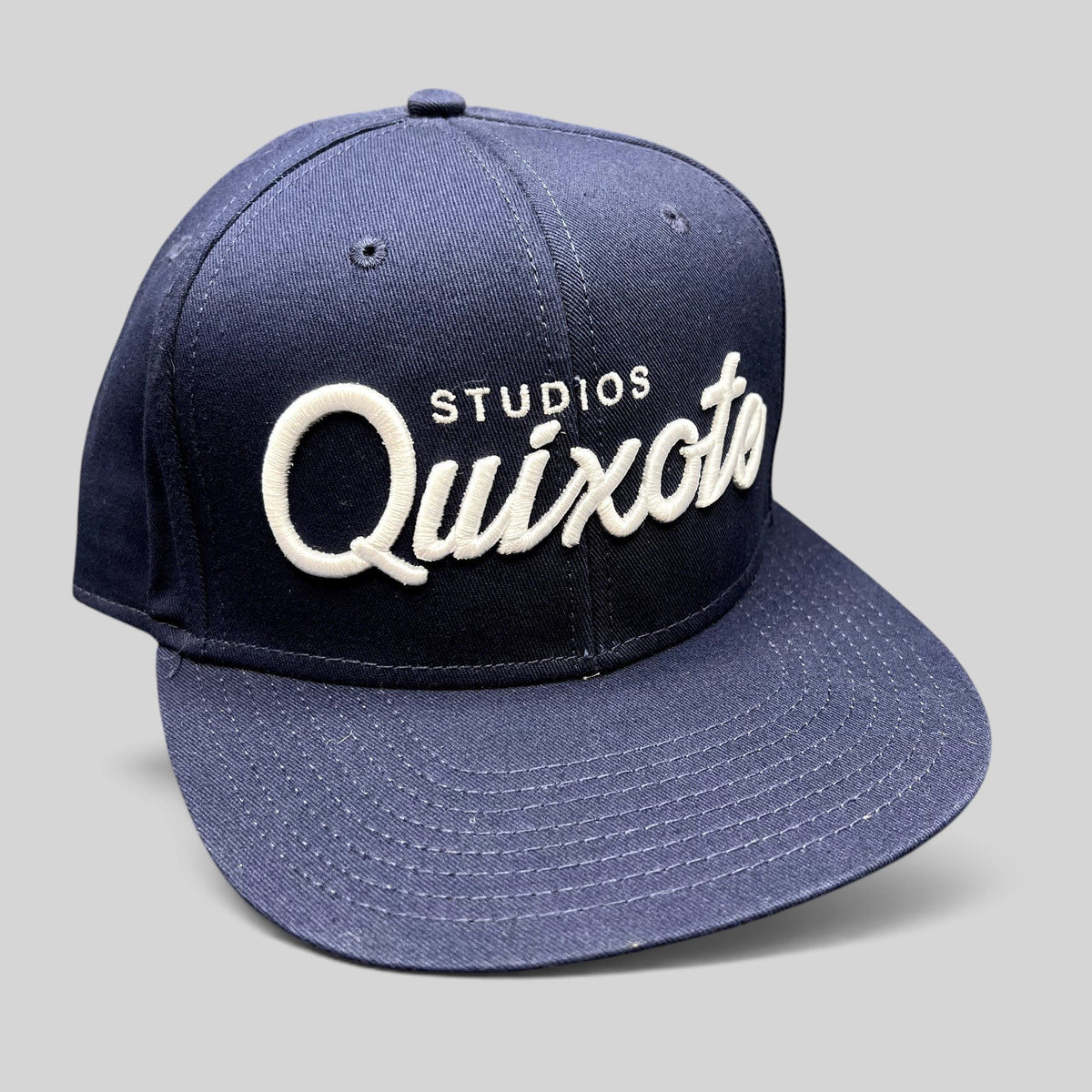 Raree Finds navy blue snapback hat with raised white Quixote Studios embroidery on the structured crown and flat brim.
