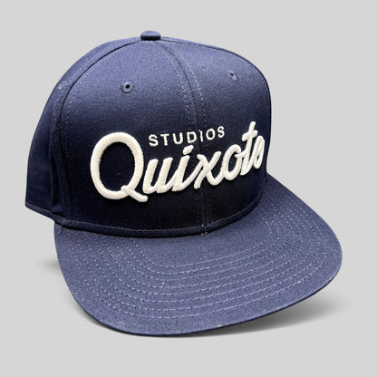 Raree Finds navy blue snapback hat with raised white Quixote Studios embroidery on the structured crown and flat brim.