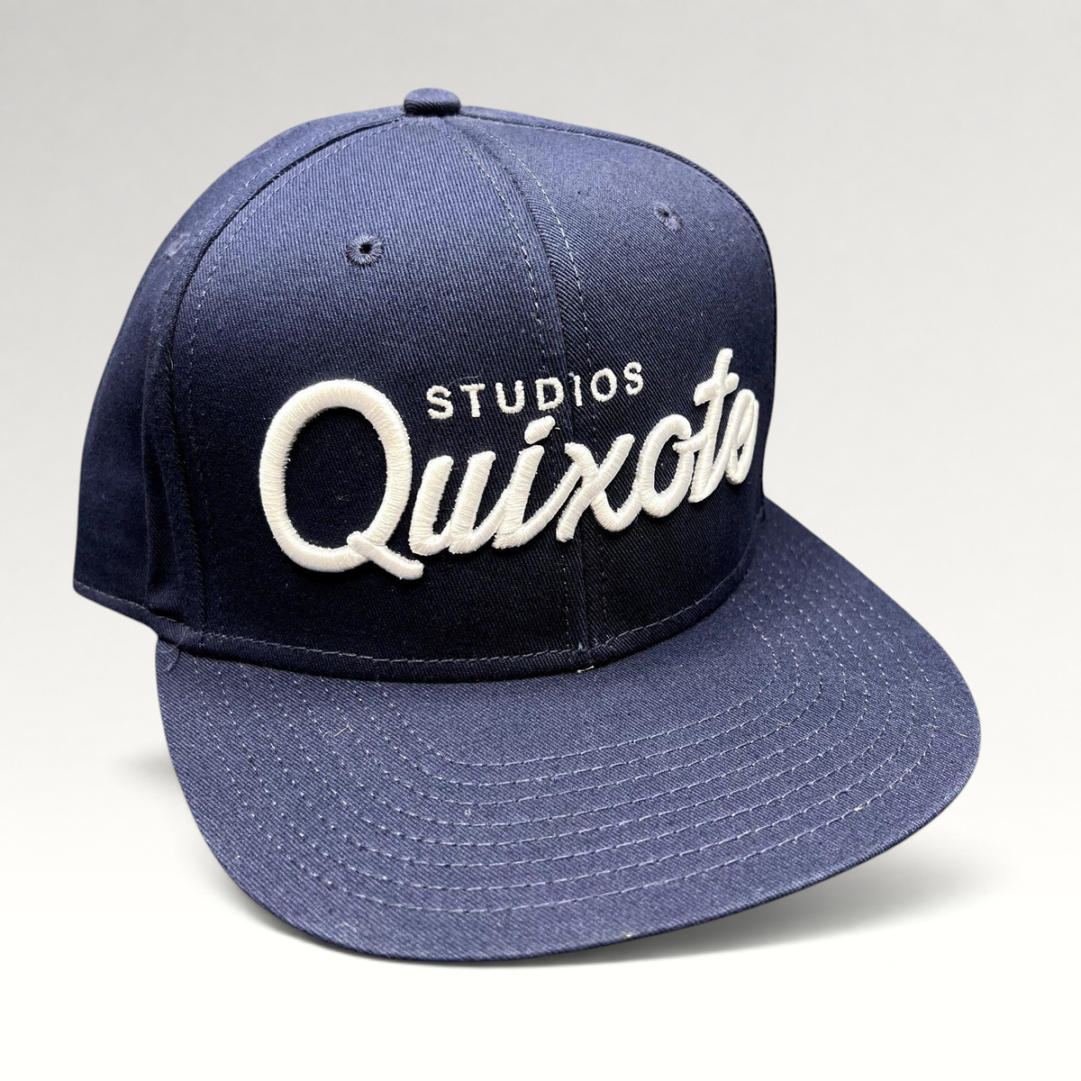 Raree Finds navy blue snapback hat with raised white Quixote Studios embroidery on the structured crown and flat brim.