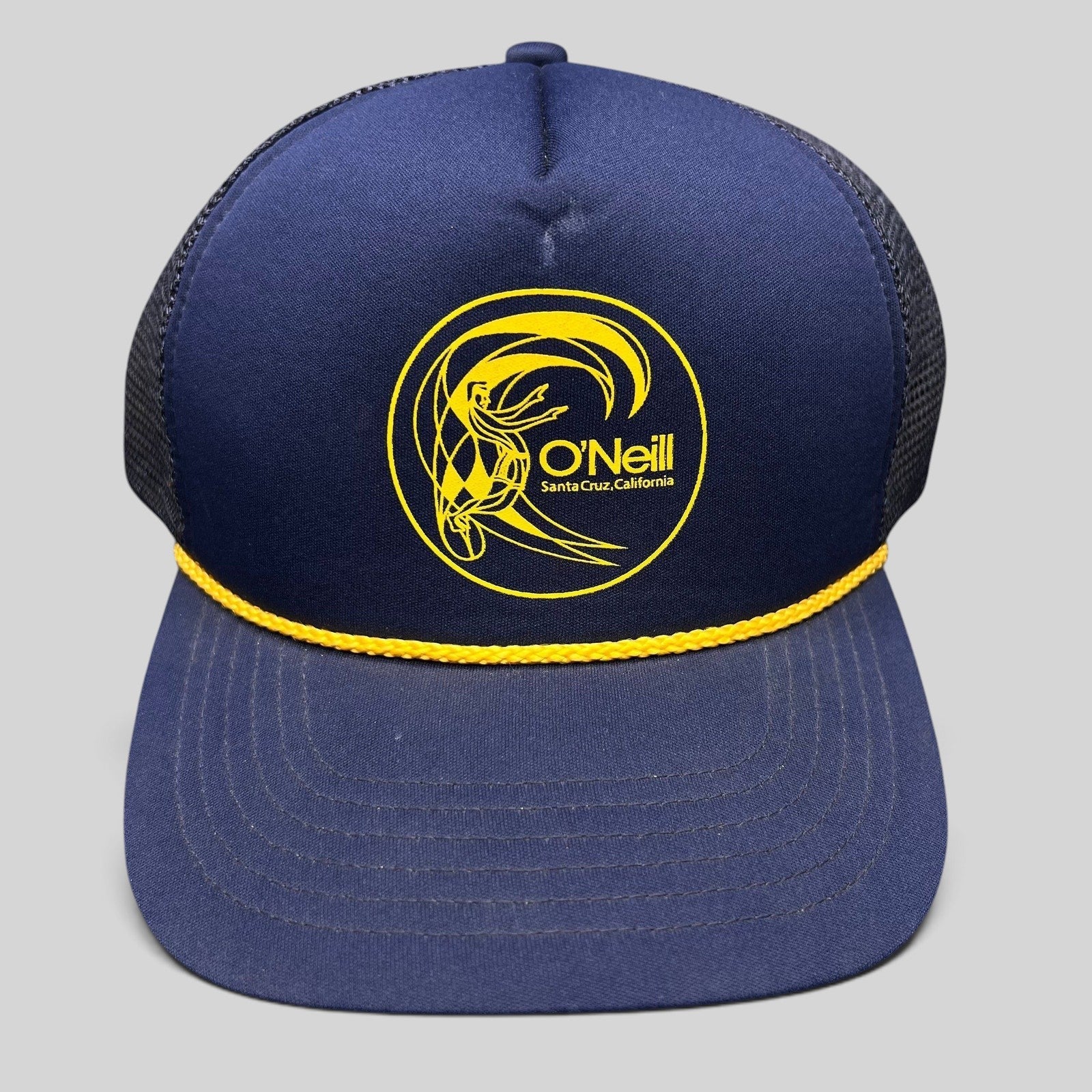 Raree Finds vintage O’Neill navy trucker hat with yellow surfer graphic, rope detail, and mesh back.
