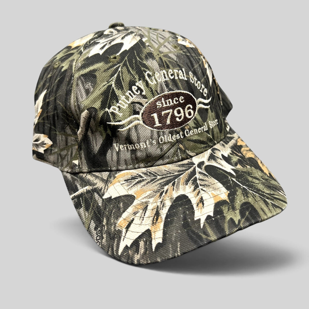 Raree Finds Putney General Store woodland camo strapback hat with embroidered 1796 Vermont logo on structured crown.