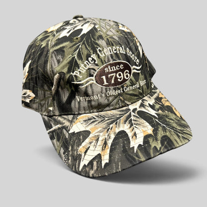 Raree Finds Putney General Store woodland camo strapback hat with embroidered 1796 Vermont logo on structured crown.