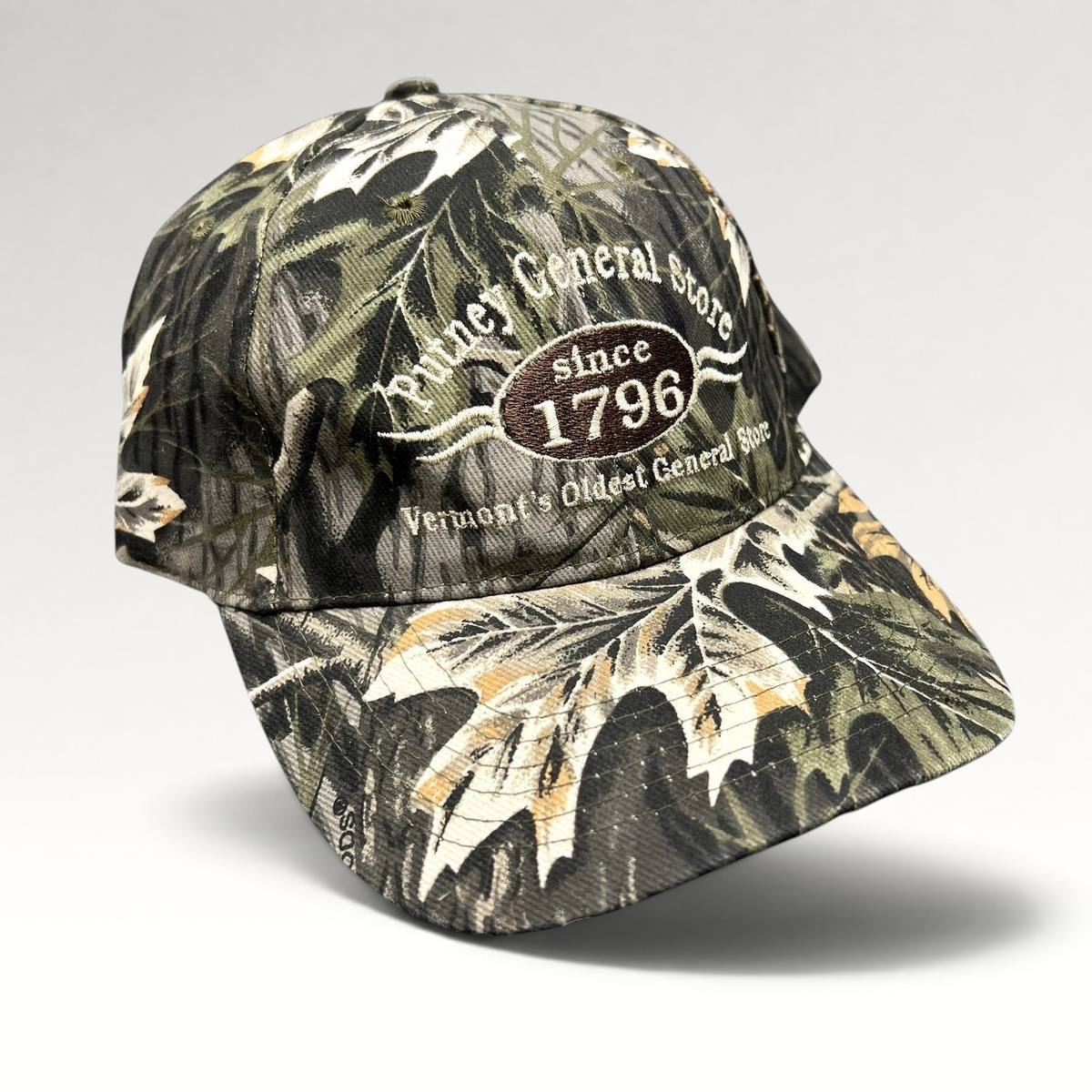 Raree Finds Putney General Store woodland camo strapback hat with embroidered 1796 Vermont logo on structured crown.