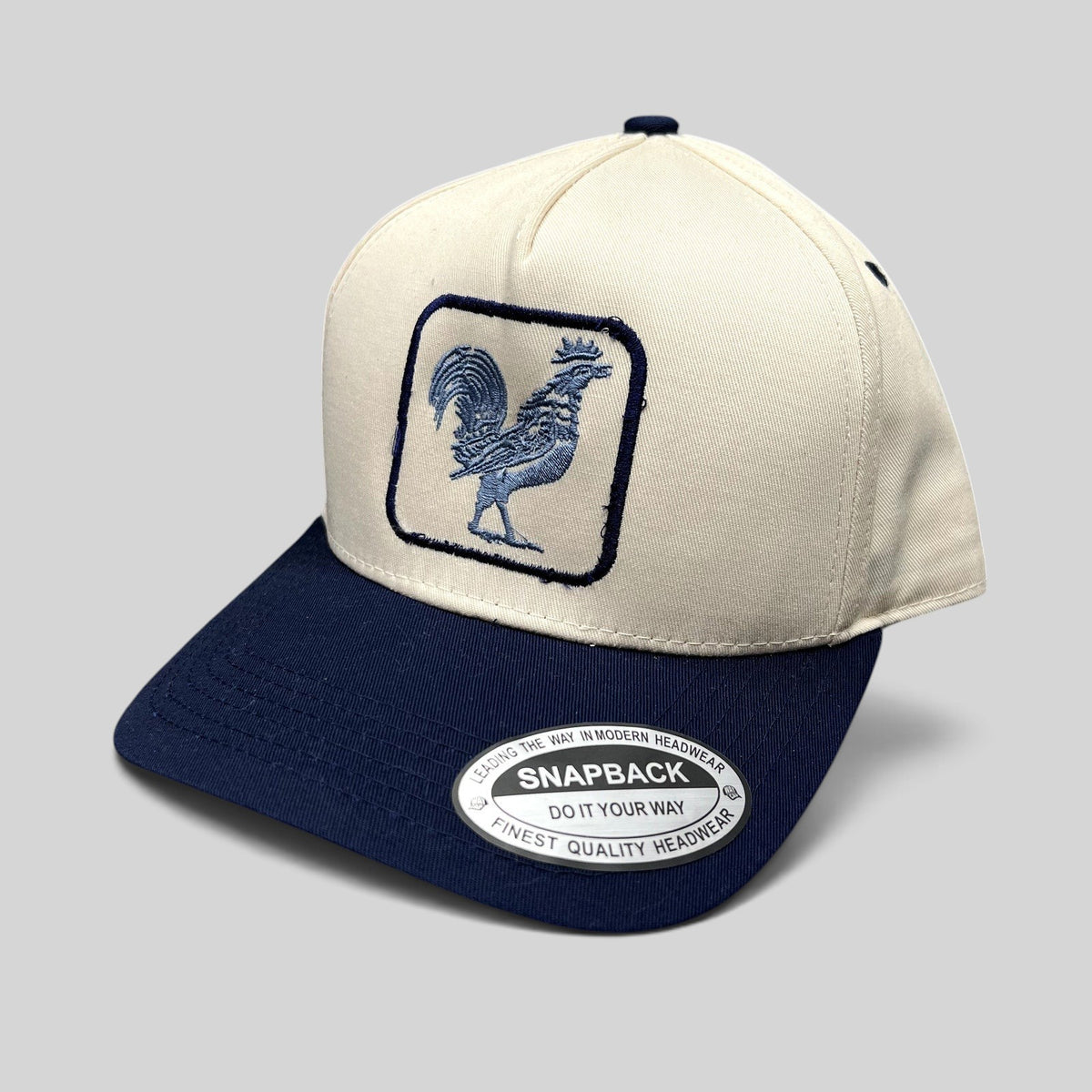 Raree Finds The Flying Cock NYC snapback hat with a cream crown, navy brim, and embroidered blue rooster front logo.