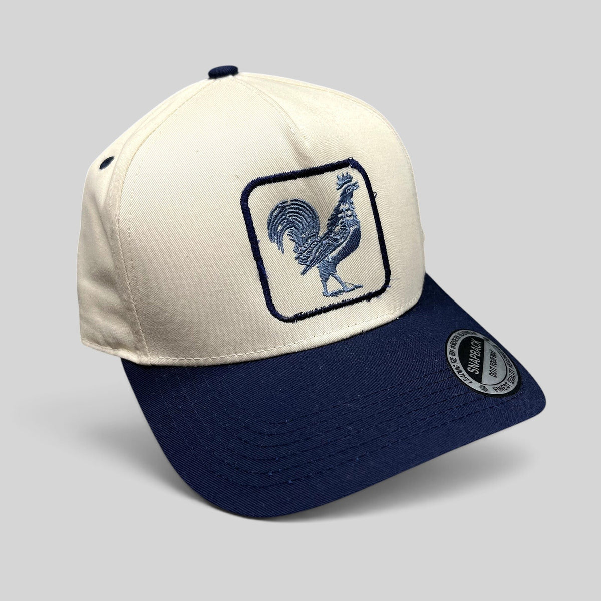 Raree Finds The Flying Cock NYC snapback hat with a cream crown, navy brim, and embroidered blue rooster logo.