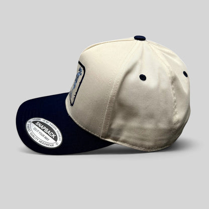 Raree Finds The Flying Cock NYC snapback hat with a cream crown, navy brim, and embroidered rooster logo side view.