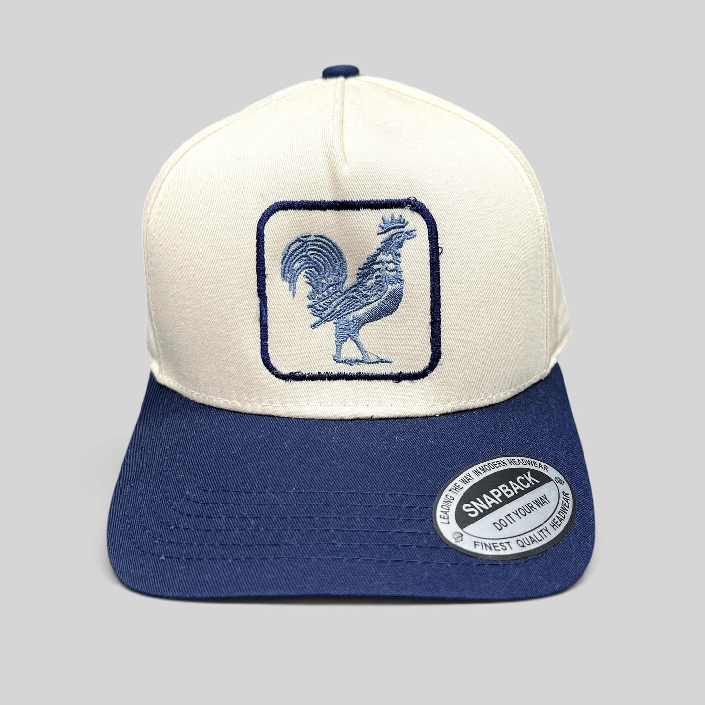 Raree Finds The Flying Cock NYC snapback hat with a cream crown, navy brim, and embroidered blue rooster front logo.