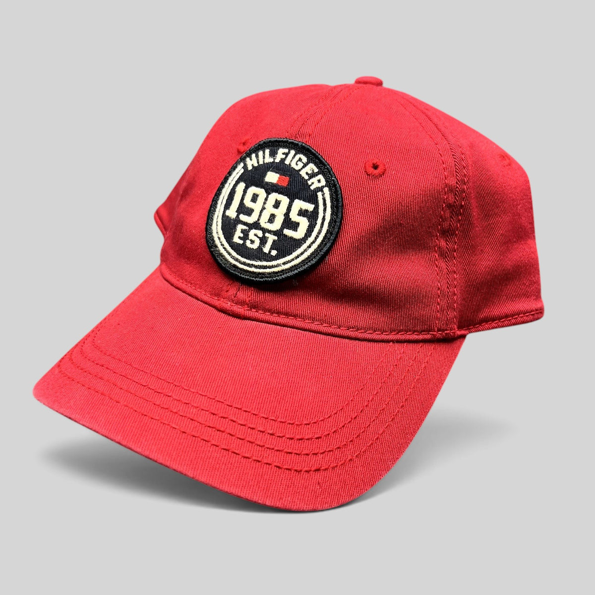Raree Finds Tommy Hilfiger red fitted hat with a circular 1985 EST patch and curved brim on a dark background.