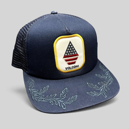 Raree Finds Volcom x New Era 9FIFTY navy trucker hat with USA flag stone logo patch and embroidered leaf brim detail.