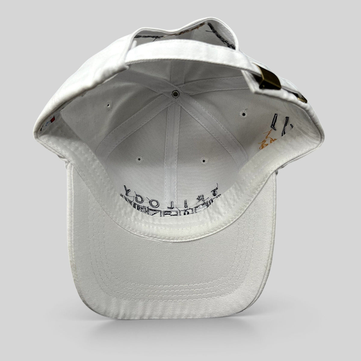 Raree Finds white cotton strapback cap with brass buckle and interior Trilogy branding, viewed from the back.