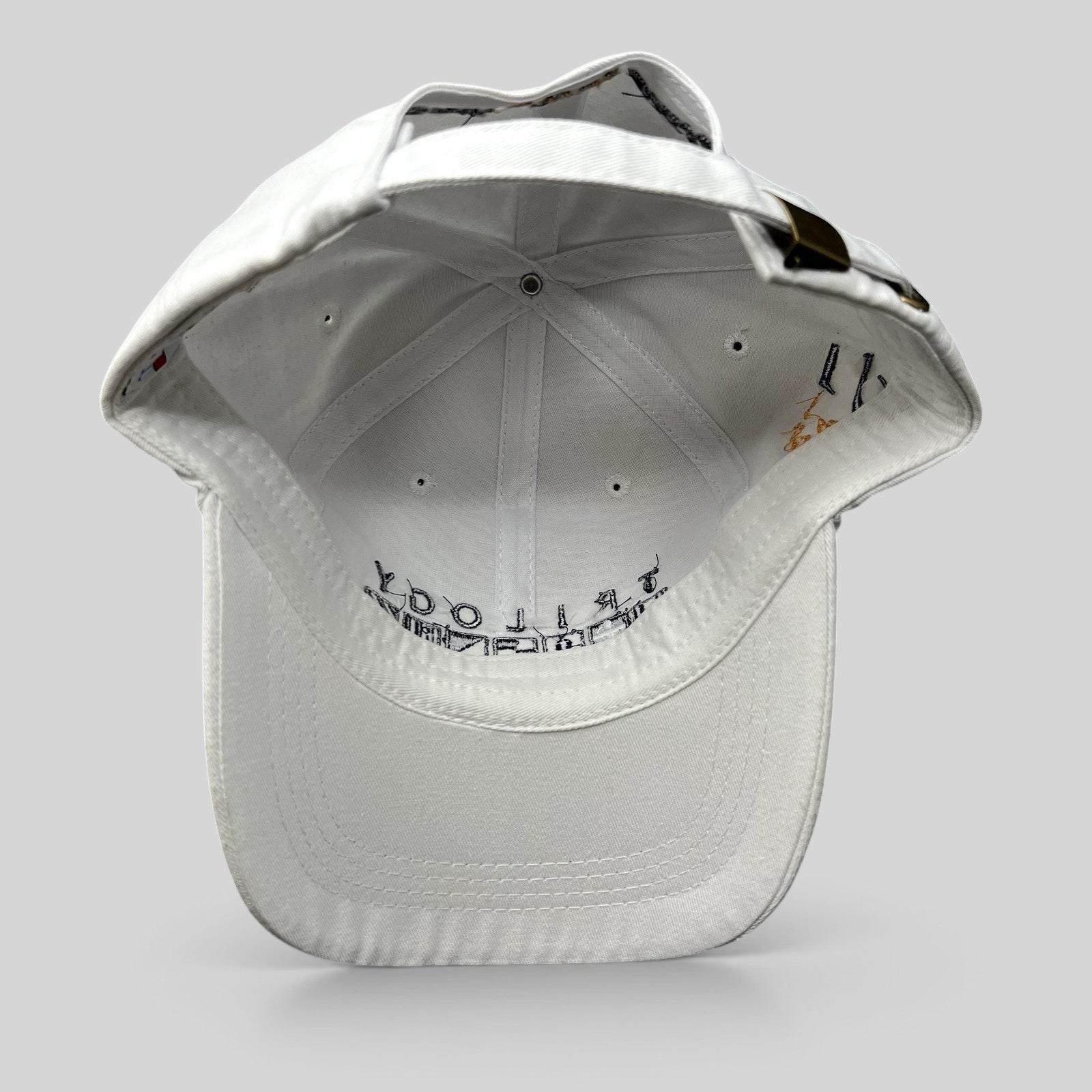 Raree Finds white cotton strapback cap with brass buckle and interior Trilogy branding, viewed from the back.