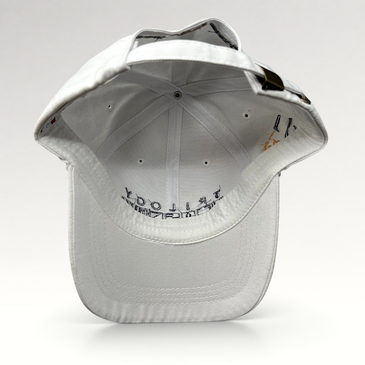 Raree Finds white cotton strapback cap with brass buckle and interior Trilogy branding, viewed from the back.