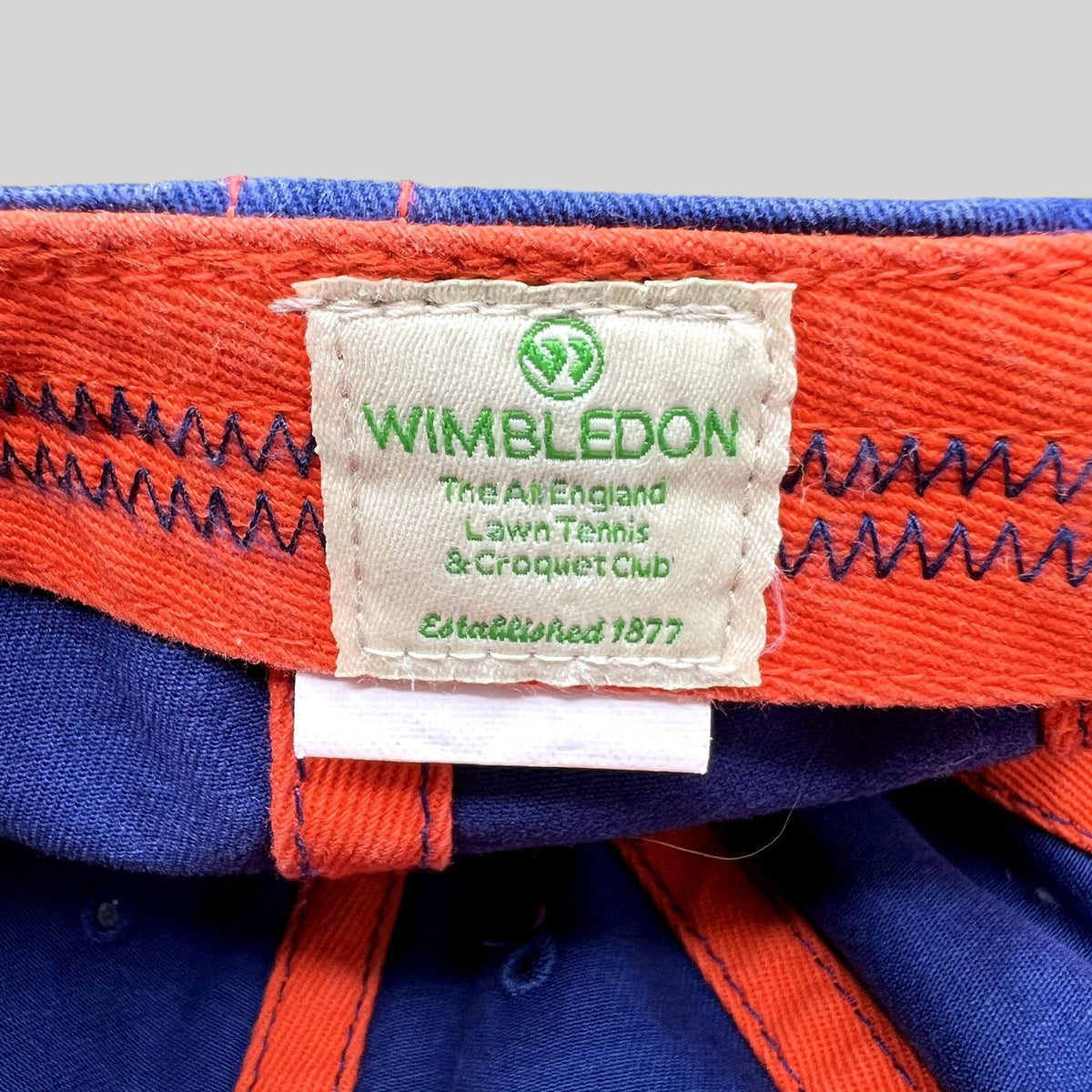 Raree Finds official Wimbledon tennis hat interior label with green logo and text on a navy and orange fabric cap