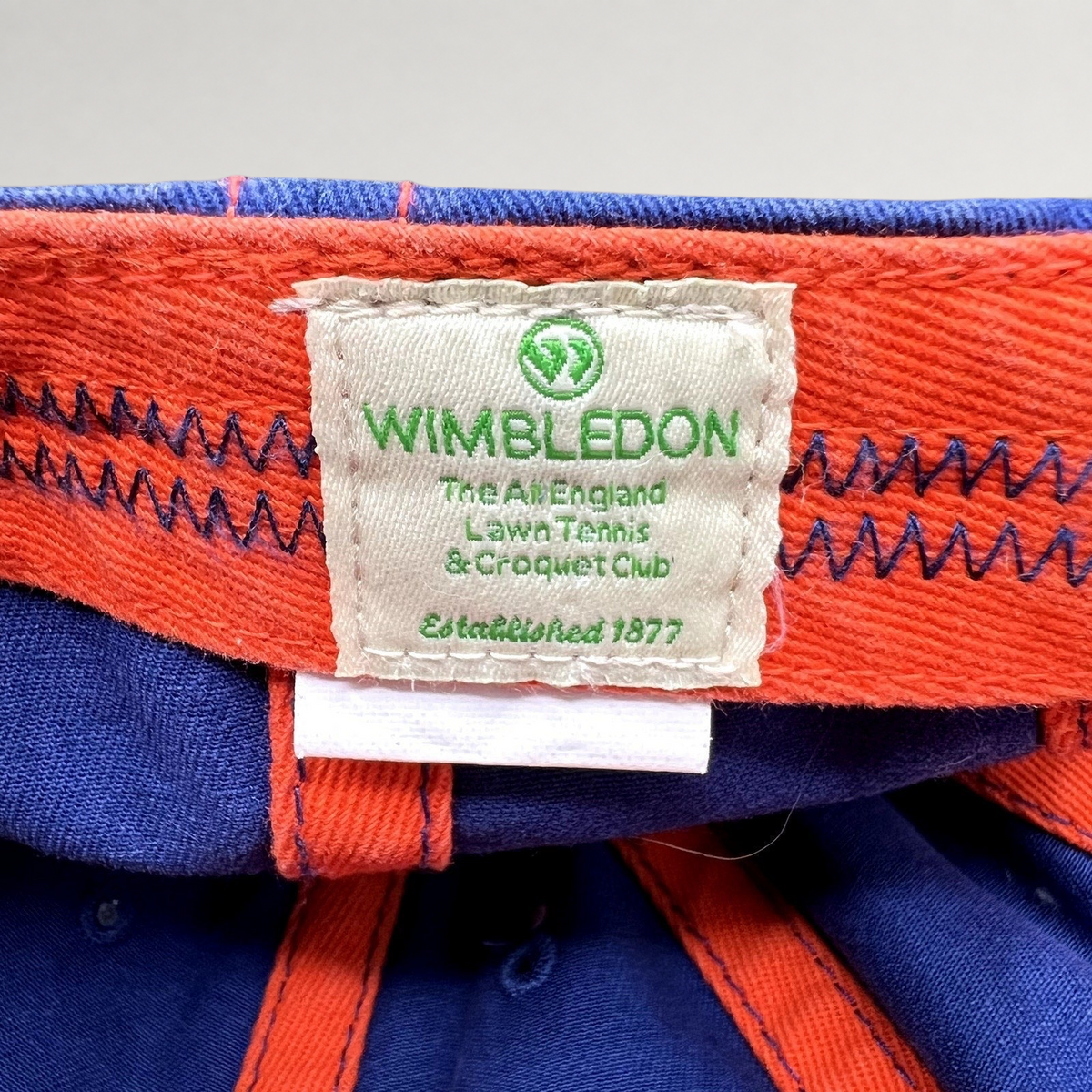 Raree Finds official Wimbledon tennis hat interior label with green logo and text on a navy and orange fabric cap